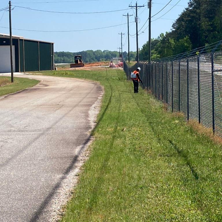 Vegetation management technician performing fence line herbicide treatment along an active airport perimeter road near secured aviation infrastructure and controlled airfield operational areas.