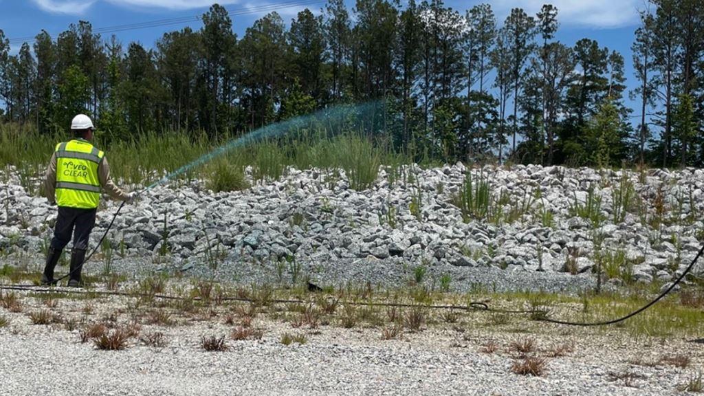 Licensed herbicide technician operating backpack sprayer on a gravel industrial site near large storage tanks, showing licensed crews performing precise industrial vegetation management and bare ground weed control at an active, regulated facility.