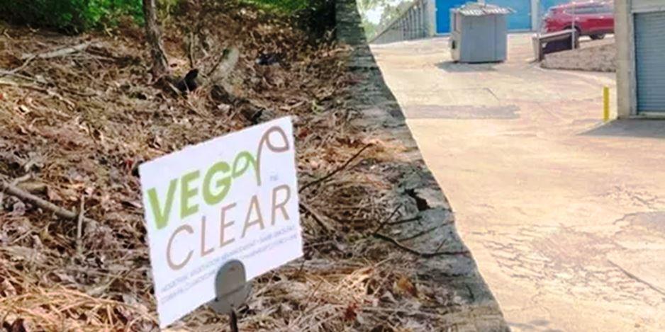 VegClear vegetation control sign displayed on a treated landfill slope next to a clean concrete access drive at a waste facility, highlighting professional herbicide application for erosion-prone embankments and critical infrastructure areas around dumpsters, loading bays, and facility entrances