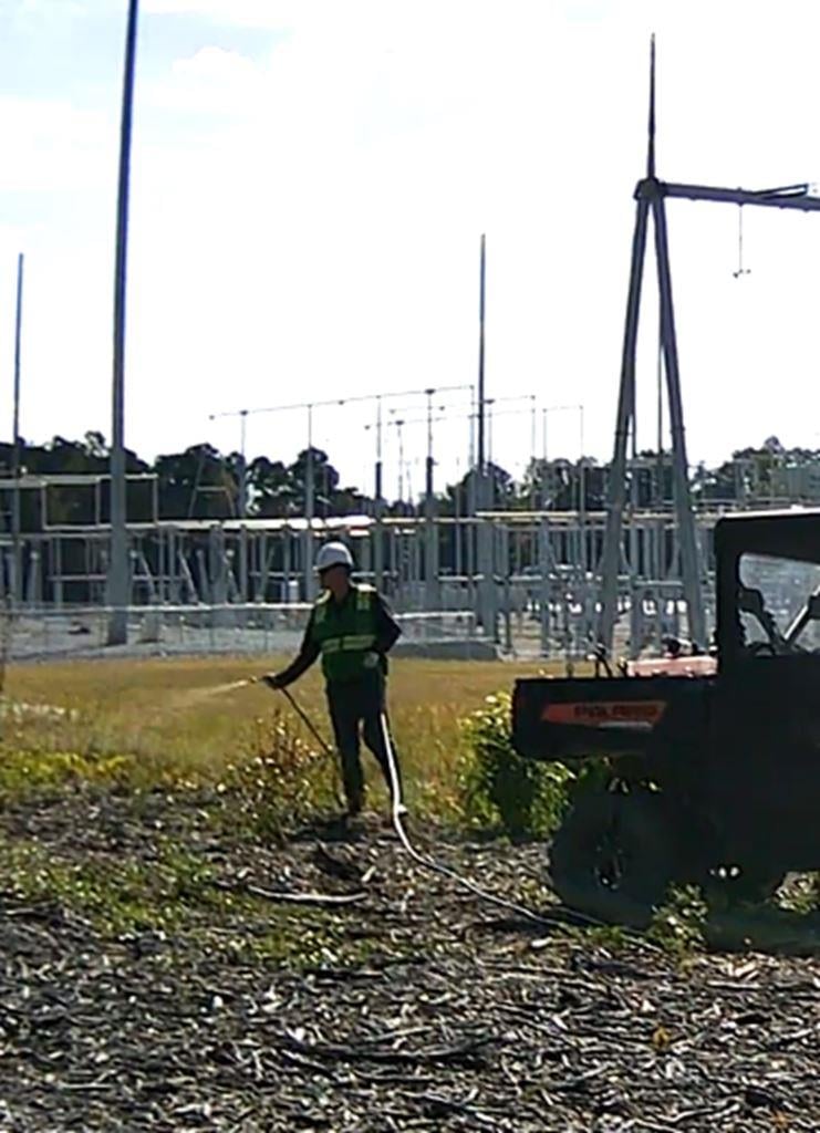 VegClear technician uses hose-fed spray system from an ATV to control brush and weeds under power lines near a substation, keeping rights-of-way clear, compliant, and safe for utility operations.