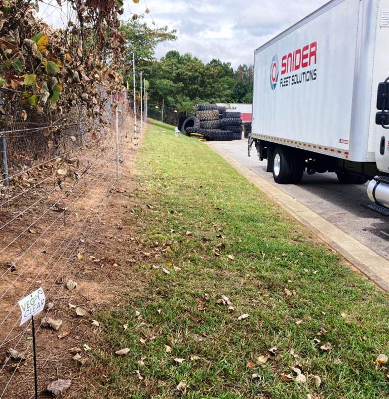 Treated fence line at Snider Fleet Solutions’ Ellenwood, GA facility following VegClear’s vegetation management service. The cleared ground shows effective herbicide application with overgrowth removed, improving visibility and security along the industrial perimeter. A Snider Fleet Solutions truck and stacked tires are visible nearby, demonstrating the active work environment and the importance of keeping fence lines free of invasive vines and brush.