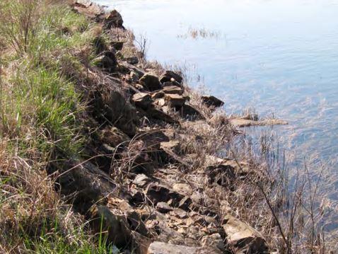 Hydroelectric dam with eroded dirt and riprap-covered embankments, requiring vegetation control strategies to prevent root intrusion and maintain safety compliance for energy production