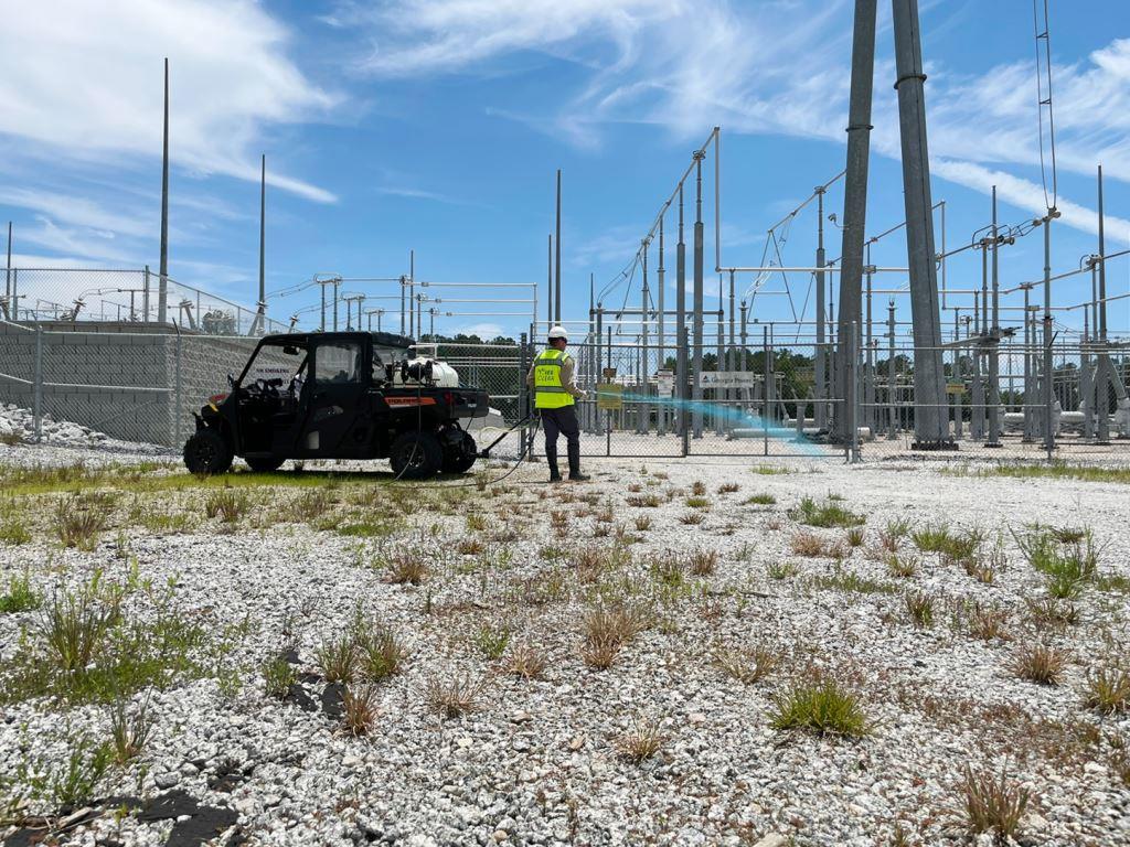 Licensed VegClear applicator performing industrial weed control at an electrical substation, applying herbicide around utility infrastructure to maintain vegetation-free gravel.