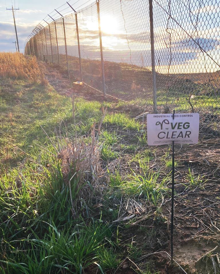 Chain link security fence with treated vegetation along the perimeter of an industrial site. Shows fence line weed control and bare ground management for access, visibility, and infrastructure protection.