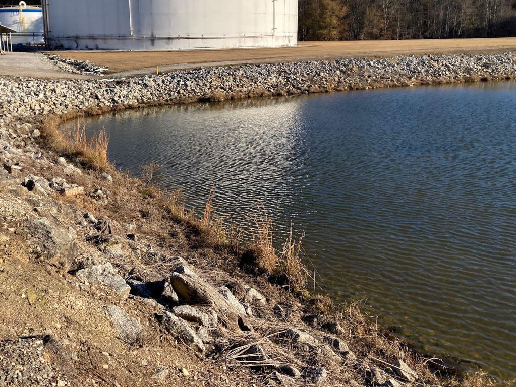 Industrial retention pond and riprap embankment adjacent to bulk storage tanks, maintained through targeted vegetation control to protect drainage, slope stability, water quality, and safe access at regulated facilities.