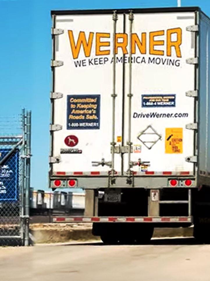 A Werner trucking rig entering an industrial outdoor storage (IOS) facility, passing through a well-maintained access road free of overgrown vegetation. This IOS yard features professionally managed weed control and herbicide-based vegetation management to ensure clear sightlines, safe truck maneuverability, and compliance with industrial property maintenance standards.