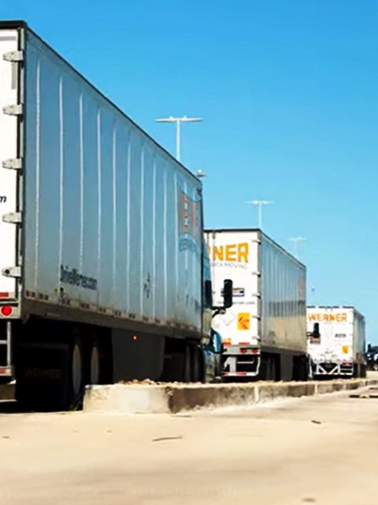Werner semi-trucks exiting an industrial outdoor storage (IOS) site with a clean, vegetation-controlled lot optimized for heavy-duty vehicle traffic. The facility's dedicated industrial weed control program prevents overgrowth along truck paths, fence lines, and loading zones, ensuring compliance with IOS property maintenance regulations and reducing safety hazards for logistics and freight operations