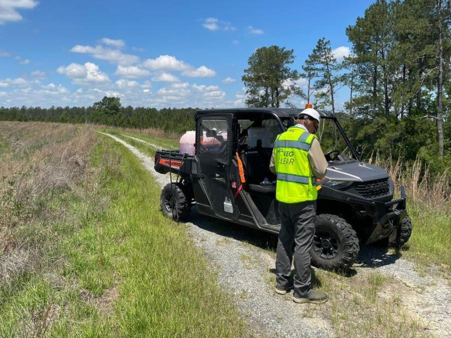 Licensed herbicide applicator prepares for industrial weed control using a UTV sprayer along an access road at a utility and infrastructure maintenance site.