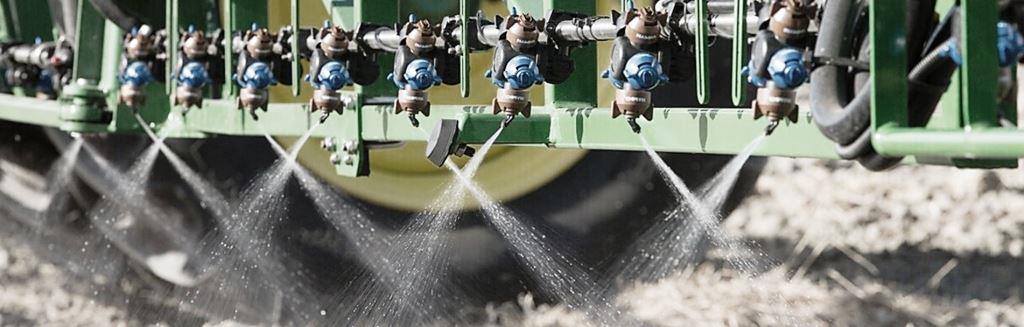 Close-up of broadcast sprayer nozzles mounted on a boom sprayer applying herbicide in uniform fan patterns for commercial vegetation management using TeeJet, Hypro, or similar spray tips.