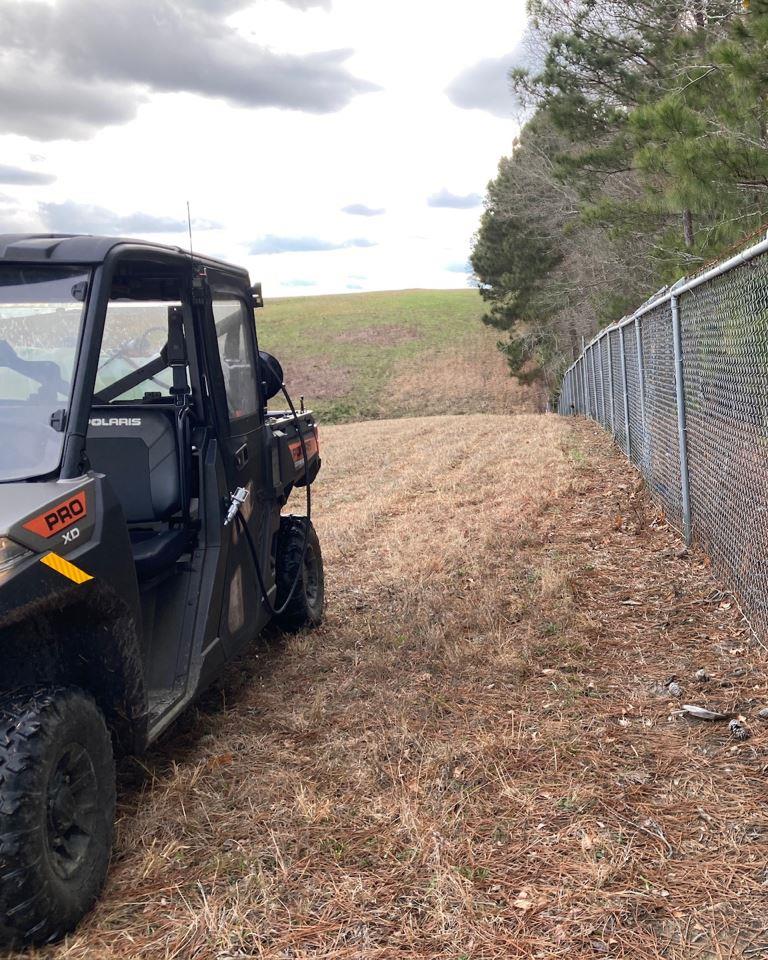 Public land vegetation management along chain link fence with utility terrain vehicle performing industrial herbicide application on grass and brush for perimeter weed control and slope maintenance.