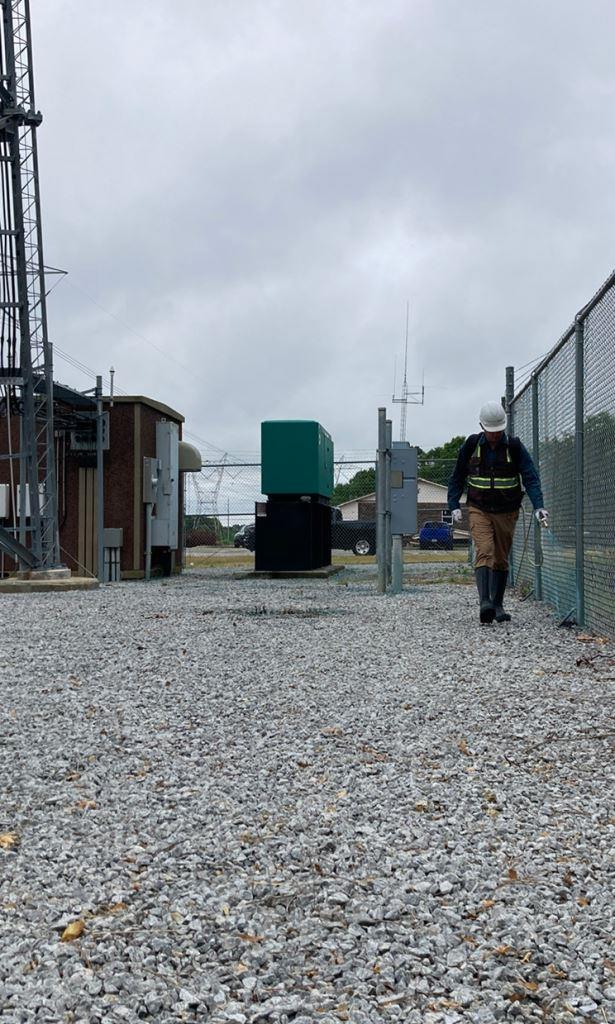 Licensed VegClear applicator treating fence line weeds in 2026 inside a secured telecom compound. Directed herbicide application along chain link fence with gravel surface, utility equipment, and controlled access site.