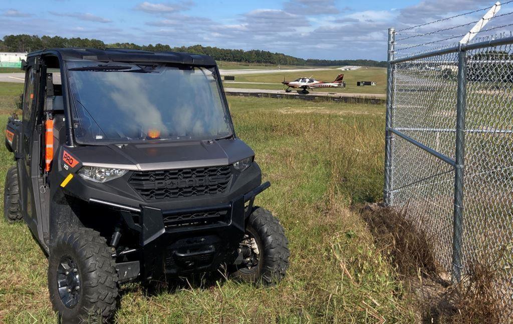 Polaris Pro XD side by side herbicide spray vehicle parked inside an airport airfield perimeter near a chain link security fence, with overgrown grass and weeds along the fence line and a small aircraft taxiing in the background on the runway.