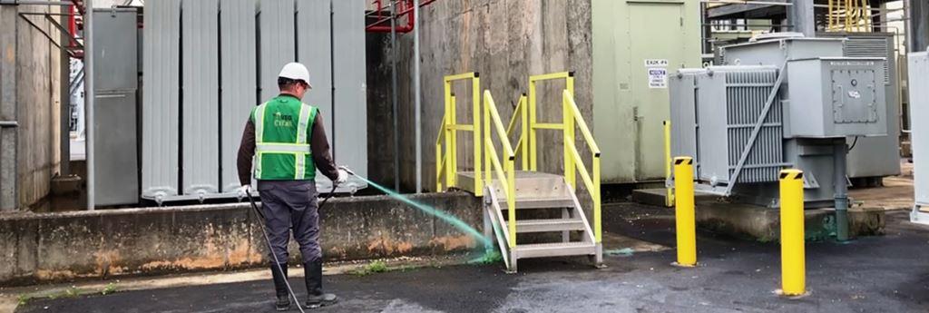 A man wearing a green VegClear hi vis safety vest and PPE is treating asphalt crack weeds with industrial herbicides in an infrastructure type environment in Georgia