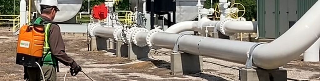 VegClear technician applying herbicide at a natural gas facility using a backpack sprayer for precision weed control around critical infrastructure, including large industrial gas pipelines and valve assemblies. Part of VegClear's industrial vegetation management services across the Southeast.