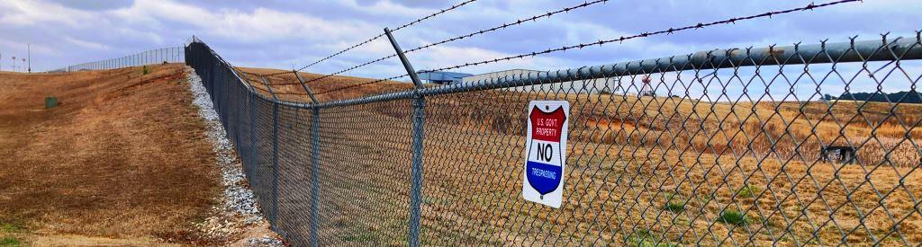 Public land perimeter fence with no trespassing sign and maintained grass slope requiring vegetation management, fence line weed control, and routine industrial grounds maintenance for security and compliance.