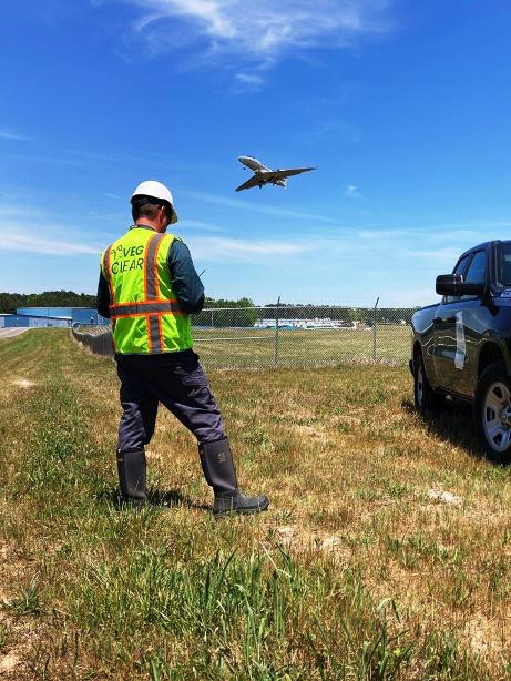 Technician maintains radio contact and inspects runway perimeter fence line vegetation at Georgia airport as aircraft departs overhead. Managed grass and weed control supports visibility, safety, and compliant airfield conditions.