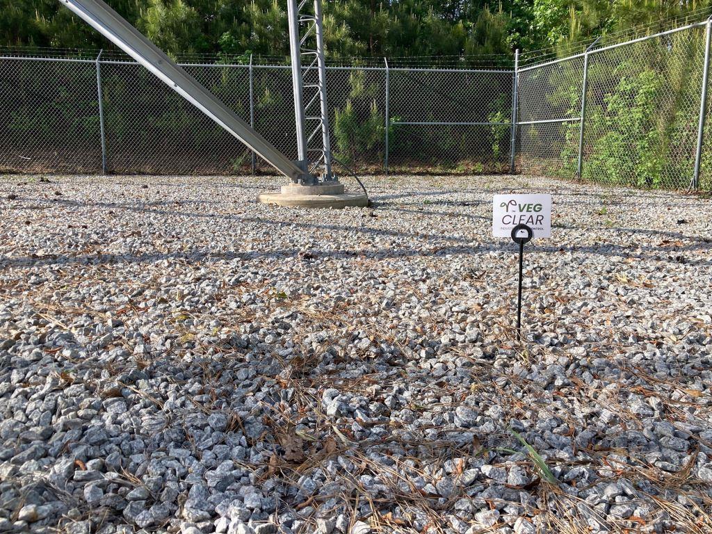 Gravel telecom cell tower compound with bare ground vegetation control, fenced perimeter, tower base foundation, and maintained utility site requiring year-round weed management.