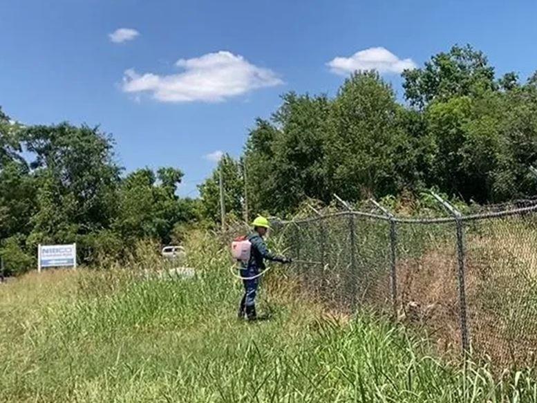 Licensed applicator using a backpack sprayer to apply selective herbicide along an industrial fence line, controlling grass and brush to maintain clear perimeters, access, and long term vegetation control