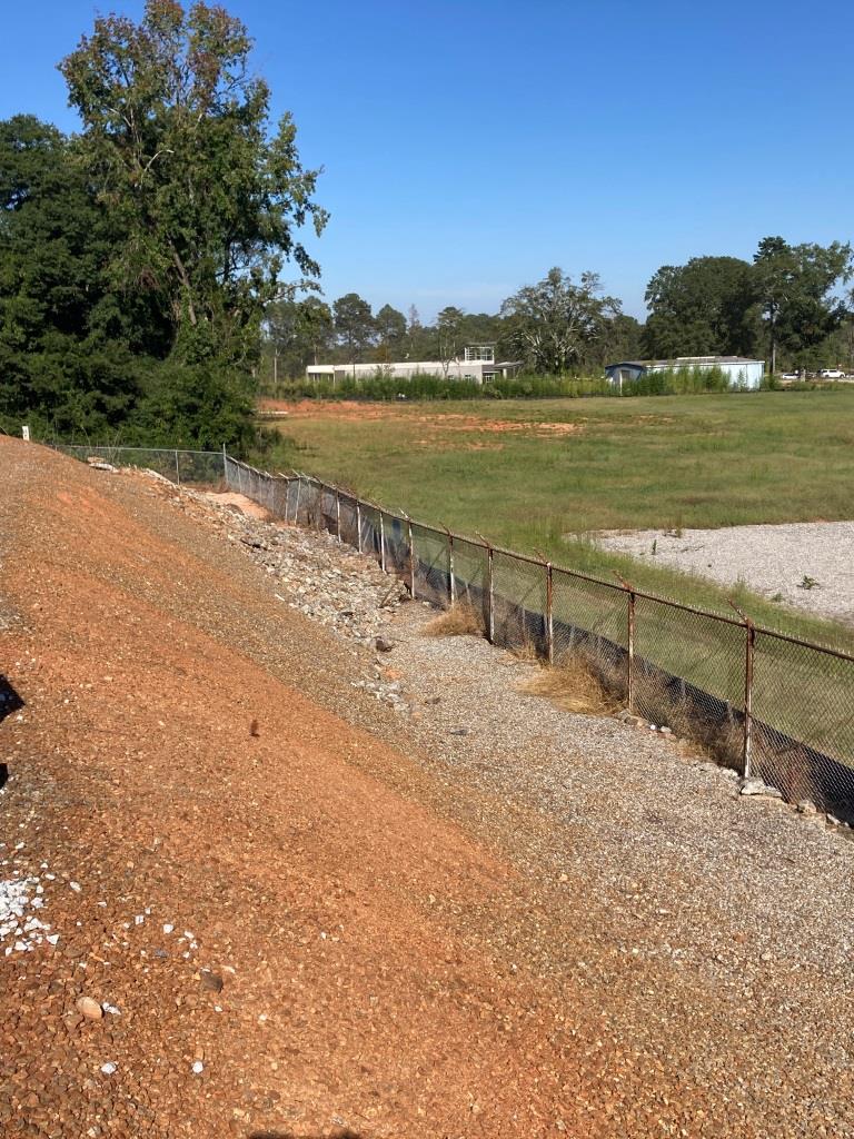 Gravel industrial embankment with treated weeds and controlled brush along a chain link fence separating a staging yard and open field, showing a managed slope with VegClear's vegetation management programs, erosion protection, and industrial weed control in Georgia
