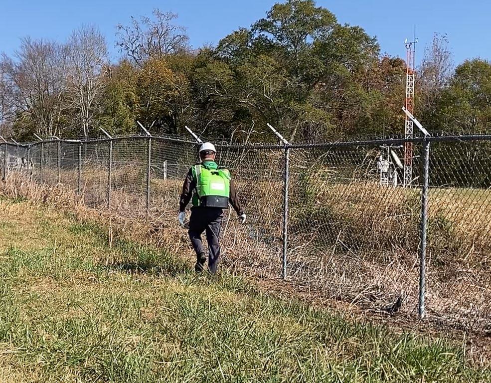 Licensed herbicide applicator performing industrial fence line weed control at a communications infrastructure site. Vegetation management treatment along a secured perimeter fence helps maintain access, visibility, security, and long-term control of grasses, brush, vines, and invasive vegetation surrounding critical utility and telecommunications assets. The herbicide application supports ongoing industrial weed control, regulatory compliance, and infrastructure maintenance programs.