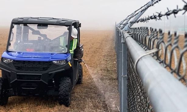 Licensed herbicide technician spraying a fence line along an industrial rail spur, applying non-selective bare ground herbicide with backpack sprayer to control weeds, grasses, and invasive vegetation. Gravel substrate treated for total vegetation control near utility infrastructure. PPE-wearing applicator following EPA-approved herbicide safety protocols during fence line weed control service at a commercial facility in Georgia.