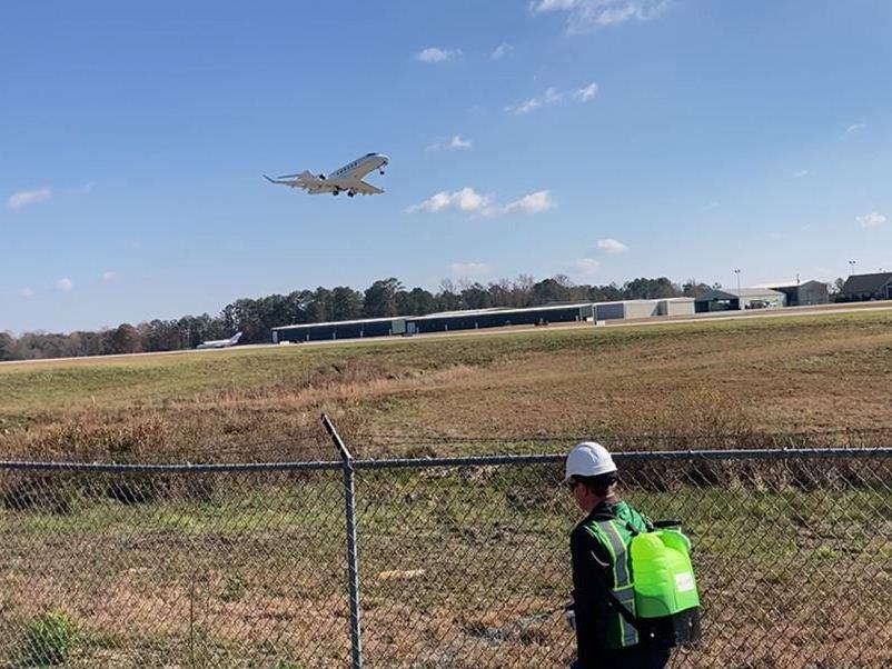 Technician applying herbicide along airport fence line while aircraft departs runway at Georgia airport perimeter vegetation control zone.