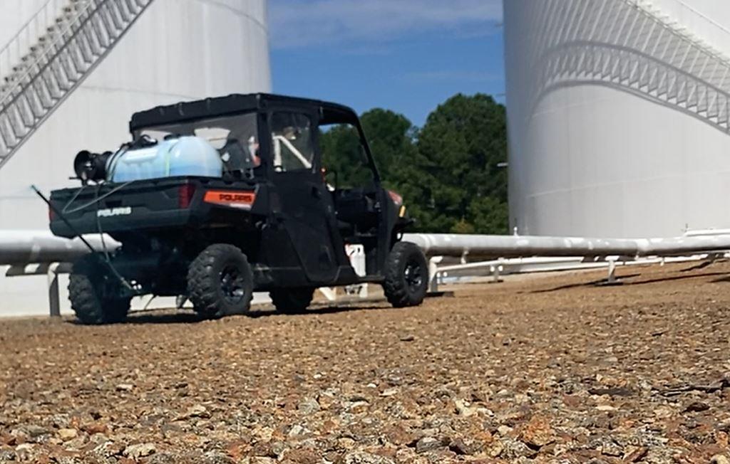 Polaris side by side with a mounted herbicide sprayer treating a gravel containment area between large fuel storage tanks. Used for industrial vegetation control to keep access routes clear, protect infrastructure, and maintain safe inspection conditions.