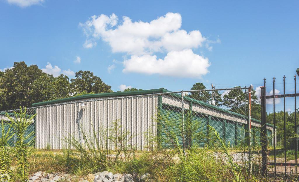 Overgrown brush and weeds along the perimeter fence of a Norcross, GA self storage facility with metal buildings and roll-up doors visible. VegClear provides fence line and brush control treatments to maintain clear, secure, and professional property boundaries.