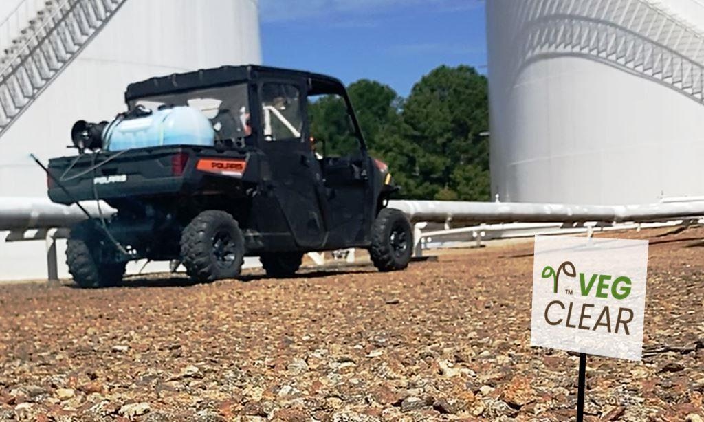 ATV mounted herbicide sprayer operating on a gravel industrial site near large storage tanks, showing licensed VegClear crews performing precise industrial vegetation management and bare ground weed control at an active, regulated facility.