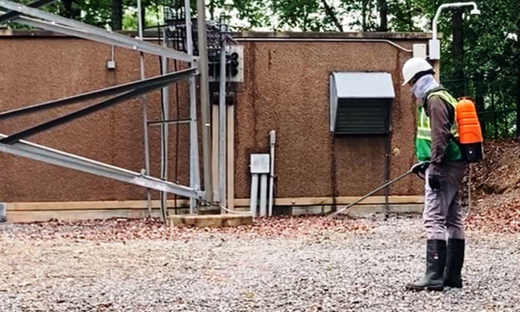 Vegetation control contractor wearing safety gear applies herbicide on gravel surface at an industrial communications site in Georgia. Image shows targeted weed management around equipment, tower legs, and utility infrastructure by a licensed technician using a backpack sprayer