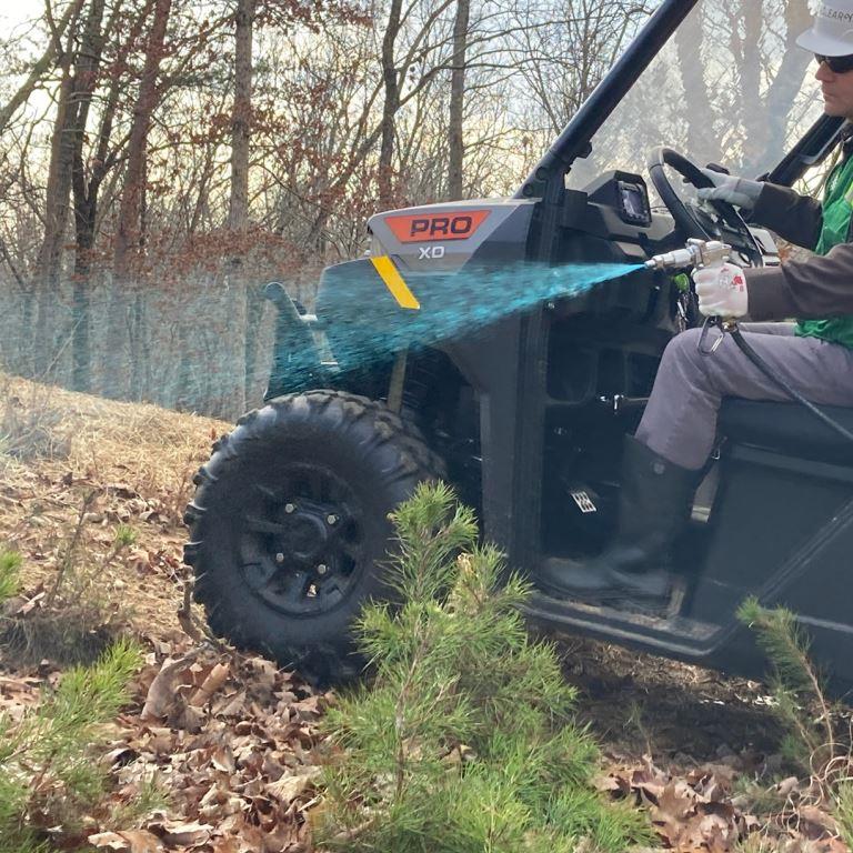 Licensed applicator performing directed winter herbicide application from a UTV to control volunteer pine saplings and woody regrowth on an industrial infrastructure site during dormant season operations