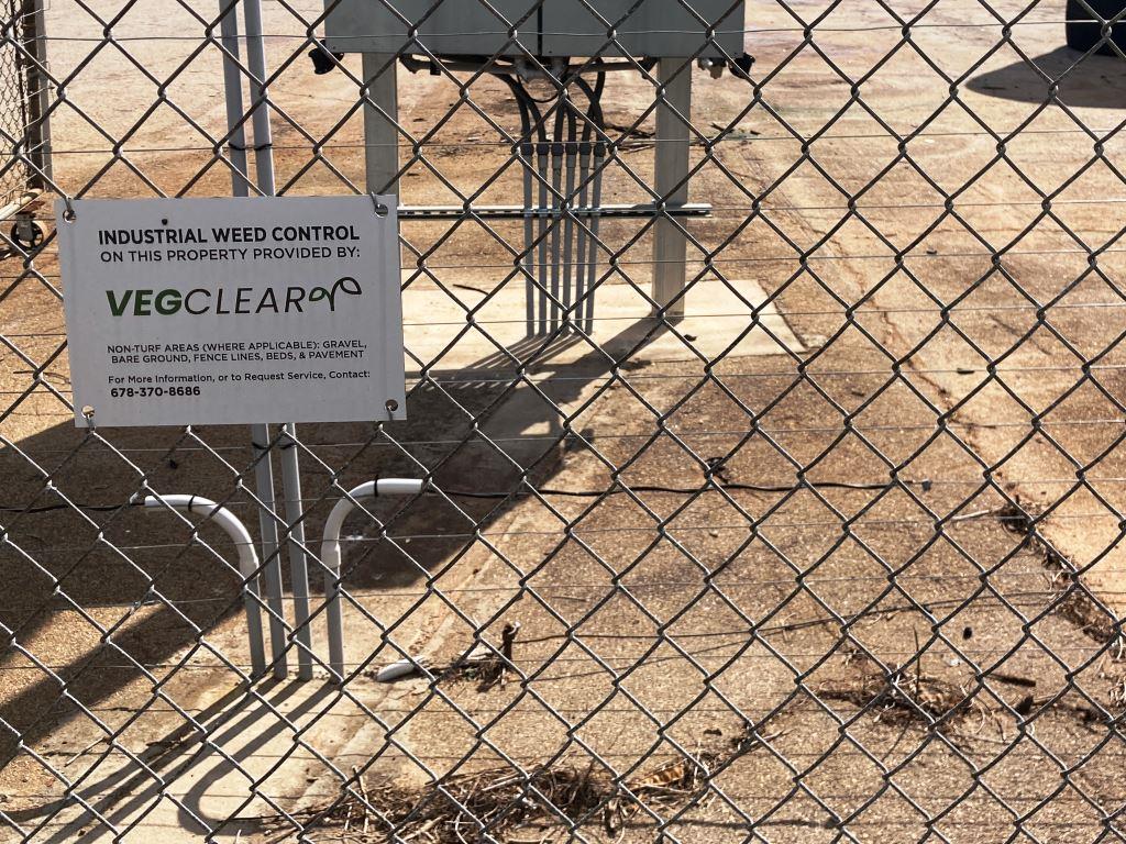 Industrial weed control signage posted on a chain link fence at a commercial logistics yard, showing VegClear responsible for vegetation management on gravel, bare ground, and fence line areas. Example site for industrial weed control case study in Georgia.