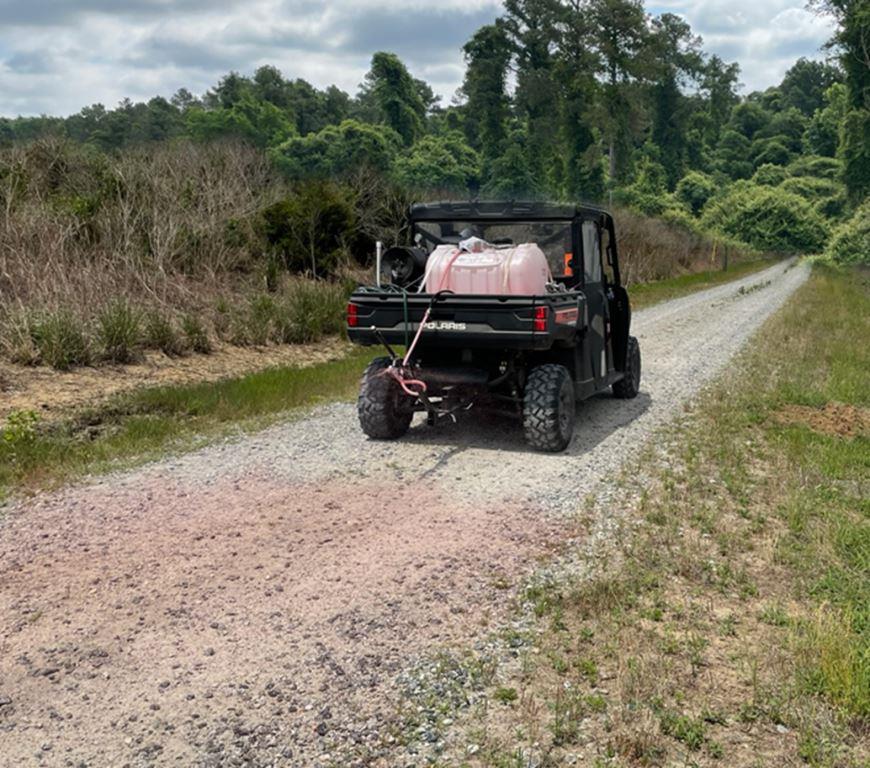 Polaris ATV sprayer applying herbicide treatment along an industrial gravel access road to control roadside vegetation and maintain clear operational access. Bare ground and right-of-way vegetation management for utility, industrial, and infrastructure sites.