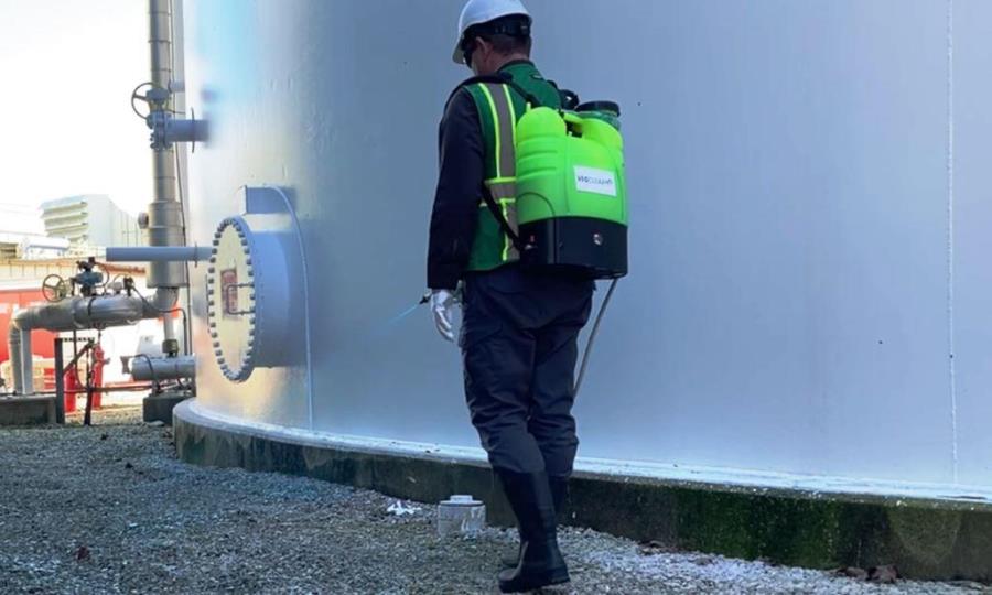 Licensed herbicide applicator performs precision weed control around industrial storage tanks using a backpack sprayer at an active manufacturing facility.
