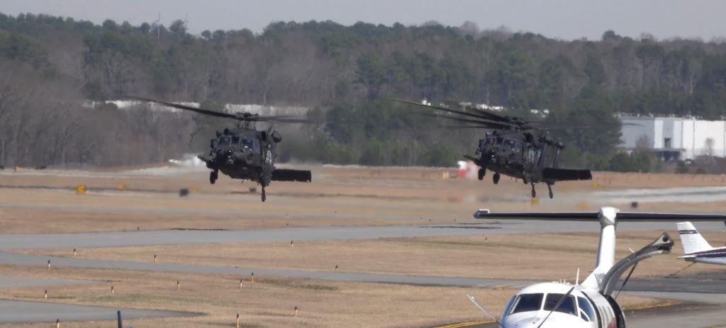 Military helicopters operating above active taxiways and maintained airfield grounds at a secured aviation facility with controlled vegetation management surrounding runways and airport infrastructure.
