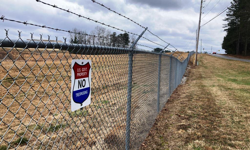 Public land security fence with no trespassing sign along roadside perimeter requiring vegetation management, fence line weed control, and routine industrial maintenance for compliance and access.