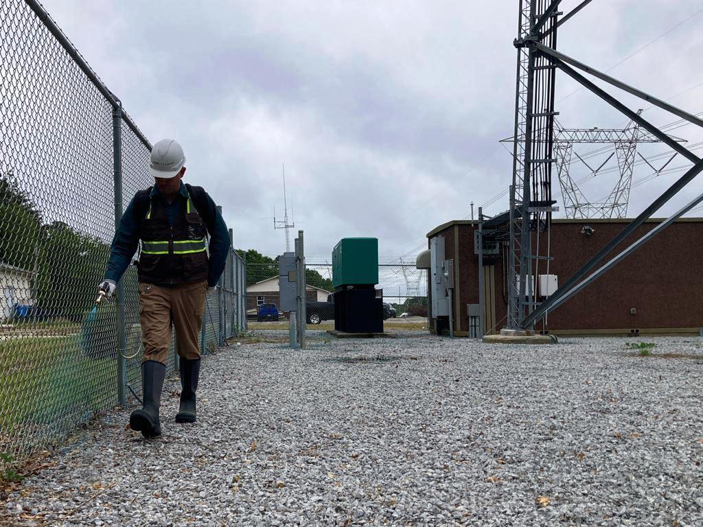 Licensed applicator performing fence line weed control at a secured utility site. Directed herbicide treatment along chain link fence beside gravel yard, utility structures, and communications equipment compound.